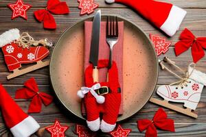 Holiday composition of plate and flatware decorated with Santa hat and clothes on wooden background. Top view of Christmas decorations. Festive time concept photo