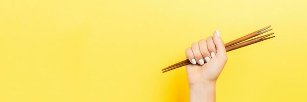 Cropped image of female hand holding chopsticks in fist on yellow background. Asian food concept with copy space photo