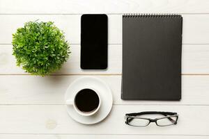 moderno oficina escritorio mesa con computadora portátil, teléfono inteligente y otro suministros con taza de café. blanco cuaderno página para usted diseño. parte superior vista, plano laico foto