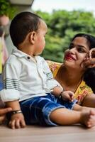 amoroso mamá que lleva de su bebé a hogar balcón. brillante retrato de contento mamá participación niño en su manos. madre con su pequeño 1 año antiguo hijo. foto