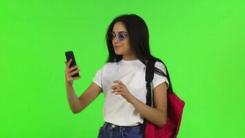 encantador fêmea aluna com uma mochila levando selfies com dela inteligente telefone video