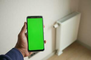 holding smart phone with green screen against White radiator on grey white wall photo