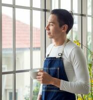 Gardener adult young Asian man one person handsome cool standing think look and Smile  relax happy drinking a cup of coffee in the room cafe with glass window and green trees in spring morning time. photo