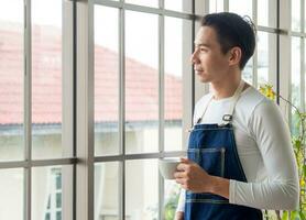 Gardener adult young Asian man one person handsome cool standing think look and Smile  relax happy drinking a cup of coffee in the room cafe with glass window and green trees in spring morning time. photo