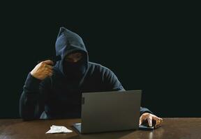 Portrait hacker spy man one person in black hoodie sitting on table looking computer laptop used login password attack security to data digital internet network system night dark background copy space photo