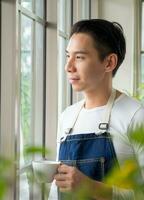 Gardener adult young Asian man one person handsome cool standing think look and Smile  relax happy drinking a cup of coffee in the room cafe with glass window and green trees in spring morning time. photo