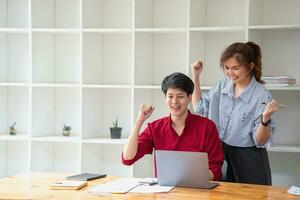 Cheerful business people using a laptop in an office. Happy young entrepreneurs smiling while working together in a modern workspace. Two young business people are sitting together at a table. photo