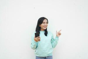 Portrait Indonesian beautiful woman wearing light blue sweater pointing to white background hold mobile phone with happy face photo