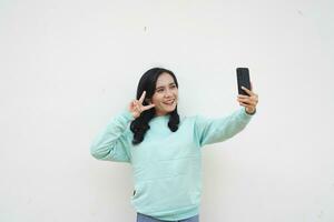Portrait Indonesian beautiful woman wearing light blue sweater take selfie with smart phone isolated on white background photo