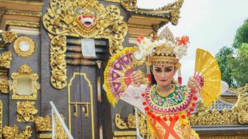 girl wearing Balinese traditional dress with a dancing gesture on Balinese temple background with hand-held fan, crown, jewelry, and gold ornament accessories. Balinese dancer woman portrait photo