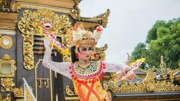 girl wearing Balinese traditional dress with a dancing gesture on Balinese temple background with hand-held fan, crown, jewelry, and gold ornament accessories. Balinese dancer woman portrait photo