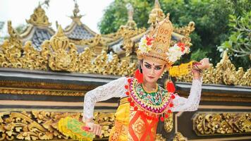 girl wearing Balinese traditional dress with a dancing gesture on Balinese temple background with hand-held fan, crown, jewelry, and gold ornament accessories. Balinese dancer woman portrait photo