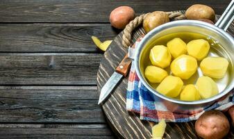 Peeled potatoes in a saucepan with fresh potatoes on the tray with a knife. photo