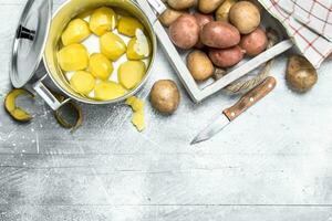 Peeled potatoes in a pot and raw potatoes on the tray. photo