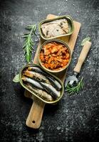 Open tin cans of canned fish on a cutting Board with a opener and rosemary. photo