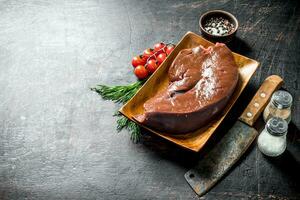 Piece of raw liver on a plate with a knife, spices and tomatoes on a branch. photo