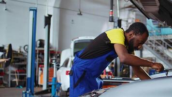 Certified engineer in car service picks torque wrench from work station bench, using it to tighten bolts after replacing engine. African american garage worker fixing client automobile video