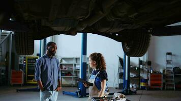 Qualified engineer in repair shop showing customer what needs to be changed on his car. Garage employee inspecting vehicle placed on overhead lift, talking with african american client video