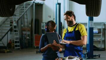 Mechanic in garage workspace standing with customer underneath suspended car, looking together for replacement parts. Employee helps african american woman fix vehicle, using laptop to find components video