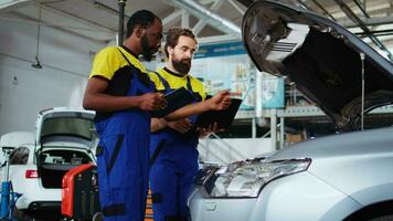 mecánica en auto reparar tienda trabajando juntos en fijación auto, utilizando tableta y ordenador portátil. compañeros de trabajo colaborando en servicio roto vehículo, comprobación para defectuoso componentes utilizando electrónico dispositivos video