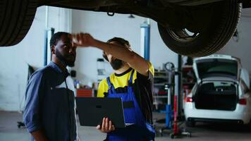 Trained mechanic and customer standing underneath car in garage using laptop to order new parts after finding defects. Employee assisting african american client by looking for components online video