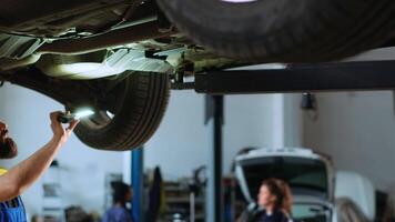 Certified repairman underneath vehicle on overhead lift in garage, checking parts. Specialist using work light to make sure automotive underbody is in proper condition, doing close examination video