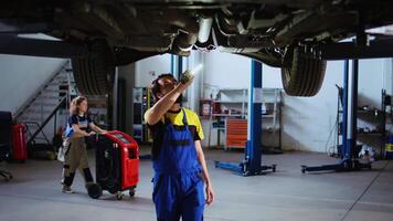 exakt serviceman arbetssätt på suspenderad bil i garage, kontroll delar under rutin- underhåll. expert- i bil reparera affär gående under fordon, läser in den använder sig av arbete ljus video