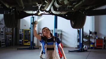 Qualified engineer working on suspended car in garage, checking components during routine maintenance. Auto repair shop employee underneath vehicle using work light to look for potential issues video