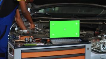 Close up shot of laptop placed on working bench in busy garage next to professional tools while serviceman fixes vehicle. Mockup device in auto repair shop with mechanic working in blurry background video