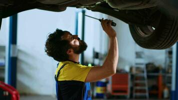 Trained serviceman underneath vehicle on overhead lift in garage, checking parts. Professional using work light to make sure automotive underbody is in proper condition, doing close inspection video