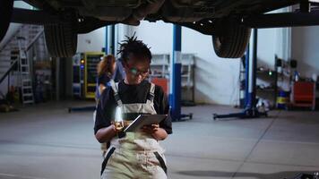 gekwalificeerd ingenieur onder geschorst auto in reparatie winkel, gebruik makend van werk licht naar controleren voor schade gedurende onderhoud. deskundige gebruik makend van professioneel gereedschap en tablet naar onderzoeken voertuig Aan overhead optillen video