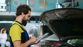 Portrait of experienced technician in auto repair shop doing car annual checkup using tablet, looking for damages. Repairman at work checking to see if vehicle components need to be changed, close up video