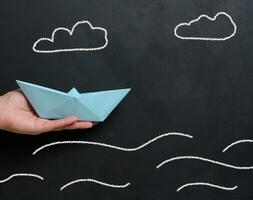 A female hand holds a paper boat against a backdrop of a board with drawn waves photo