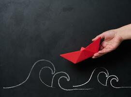 A female hand holds a red paper boat, with chalk-drawn waves below, representing the concept of mentorship and business support photo