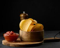 Potato chips in a wooden bowl and a bowl of sauce on a black table, snack photo