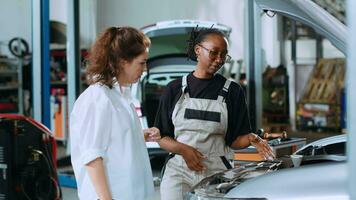 simpático mecánico ayudando cliente con coche mantenimiento en auto reparar tienda. calificado especialista en garaje mirando terminado coche componentes con mujer, servicio su vehículo durante anual chequeo video