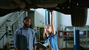 Meticulous technician in garage workplace stands with customer underneath suspended car, looking together for engine replacement. Expert helps client mend vehicle using device to find needed part video