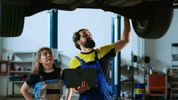 Certified serviceman in garage workspace standing with customer underneath suspended car, looking together for replacement parts. Expert helps woman fix vehicle, using laptop to find components online video