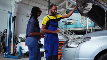 Qualified serviceman in repair shop showing customer what needs to be changed on her car for it to properly work. Garage specialist inspecting vehicle placed on overhead lift, discussing with client video