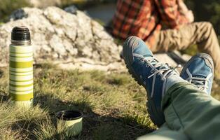 Women's legs in hiking boots relaxing on mountain near river in autumn and man in red plaid shirt looking at view and drinking tea from thermos Tourism photo