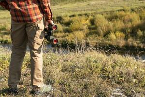 faceless man in red plaid shirt with binoculars standing on river bank near cliff in fall Birdwatching and tourism copy space photo