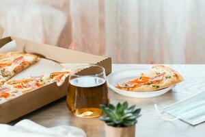 Takeout food. A slice of pizza in a disposable plastic plate and a box of pizza on the table in the kitchen. photo