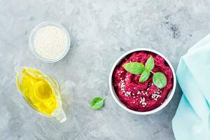 Homemade baked beetroot hummus in a bowl with sesame seeds and basil on the table. Top view photo