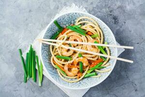 Ready to eat udon and wok noodles with prawns, peppers and onions in a plate and chopsticks on the table. Chinese food. Top view photo