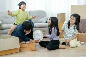 Moviente casa, reubicación familia sonrisa y jugar juntos después moverse a nuevo hogar, dentro el habitación estaba un cartulina caja Contiene personal pertenencias y mueble. moverse en el nuevo departamento, condominio foto