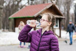 un linda niña en lentes bebidas agua desde un botella compró en un comida camión en un ciudad parque. para llevar comida foto