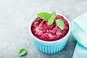 Homemade baked beetroot hummus in a bowl with sesame seeds and basil on the table. Close-up photo