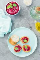 Homemade bruschetta with beetroot hummus on small baked baguette toast on a plate and a bowl of hummus on the table. Vertical and top view photo