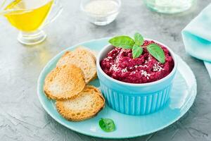 Homemade baked beetroot hummus in a bowl with sesame seeds and basil and baked small toast on a plate on the table photo