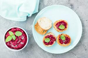 Homemade bruschetta with beetroot hummus on small baked baguette toast on a plate and a bowl of hummus on the table. Top view photo